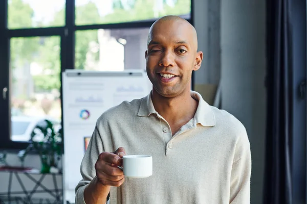 Chronic illness, inclusion, bold african american man with myasthenia gravis disease holding cup of coffee, happy and dark skinned office worker with ptosis syndrome looking at camera — Stock Photo