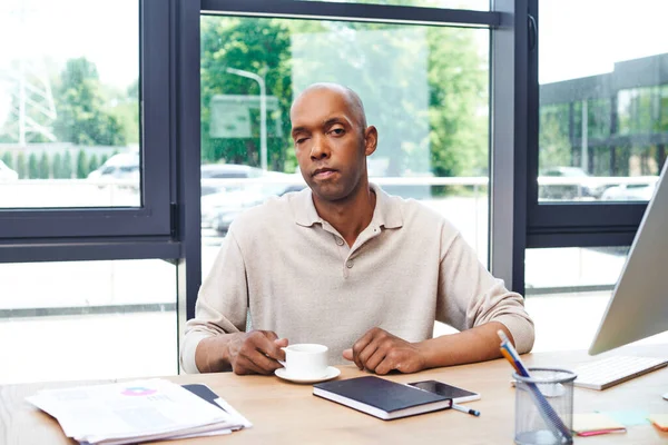 Inclusión, hombre afroamericano con miastenia grave enfermedad sentado en el escritorio con taza de café, trabajador de oficina de piel oscura en traje casual mirando a la cámara, monitor, teléfono inteligente, gráficos en la mesa - foto de stock