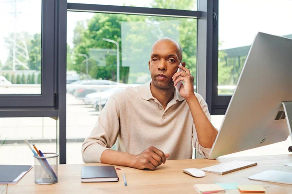 Myasthenia disease, bold african american man at work, dark skinned office worker with ptosis syndrome talking on smartphone, inclusion, corporate culture, monitor and stationery on desk — Stock Photo