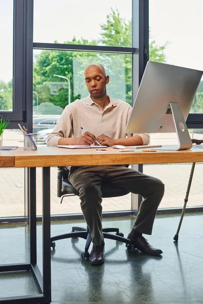 Inclusion, african american man with myasthenia at work, bold and dark skinned office worker, corporate culture, monitor and smartphone, computer mouse, keyboard and stationery on desk — Stock Photo