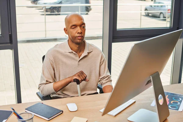 Inclusion, african american man with myasthenia gravis, bold and dark skinned office worker sitting with walking cane and using computer, looking at monitor, graphs and notebook on desk — Stock Photo