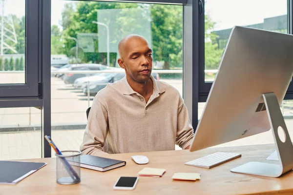 Inclusion, homme afro-américain audacieux avec myasthénie grave, employé de bureau à la peau foncée assis au bureau et utilisant un ordinateur, regardant moniteur, clavier et souris, smartphone avec écran blanc — Photo de stock