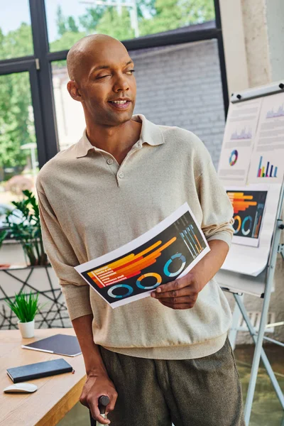 Personas reales, hombre afroamericano audaz con miastenia gravis, trabajador de oficina de piel oscura de pie con caña y gráficos de celebración, enfermedades crónicas, diversidad e inclusión — Stock Photo