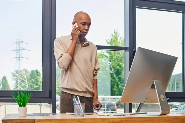 Myasthenia gravis, смелый африканский бизнесмен с синдромом птоза, разговаривающий на смартфоне и стоящий с тростью рядом с монитором, темнокожий офисный работник, включение — стоковое фото