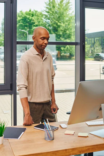 Myasthenia gravis syndrome, bold african american businessman standing with walking cane and looking at computer monitor, dark skinned office worker in casual attire standing at desk, inclusion — Stock Photo
