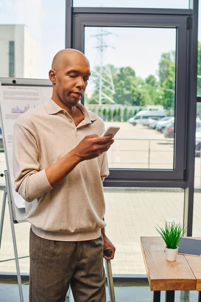 Myasthenia gravis, bold african american businessman with eye syndrome using smartphone and standing with walking cane, dark skinned office worker with ptosis syndrome near desk, inclusion — Stock Photo