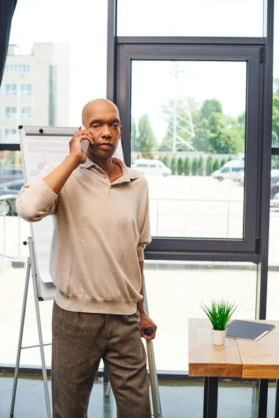 Bold african american businessman with eye syndrome talking on smartphone and standing with walking cane, dark skinned office worker with ptosis syndrome near desk, inclusion — Stock Photo
