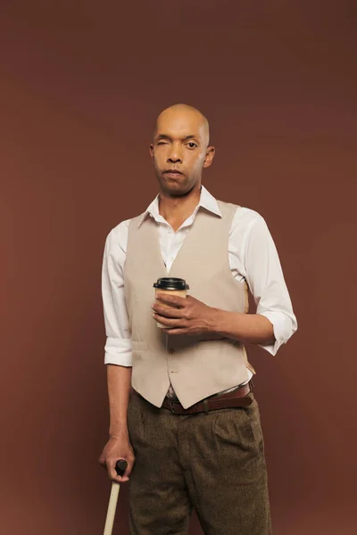 Inclusion, bold african american man with myasthenia gravis syndrome, standing with paper cup and walking cane, dark skinned man with chronic disease on brown background, coffee to go — Stock Photo