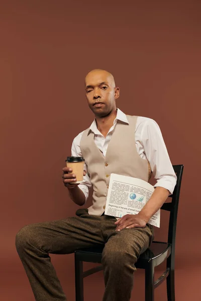 Inclusão, homem americano africano ousado com síndrome de miastenia gravis, sentado na cadeira e segurando copo de papel com jornal, café para ir, homem de pele escura com doença crônica em fundo marrom — Fotografia de Stock