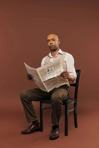Inclusão, homem americano africano ousado com síndrome da miastenia gravis, sentado na cadeira e lendo jornal, homem negro de pele escura com doença crônica em fundo marrom — Fotografia de Stock