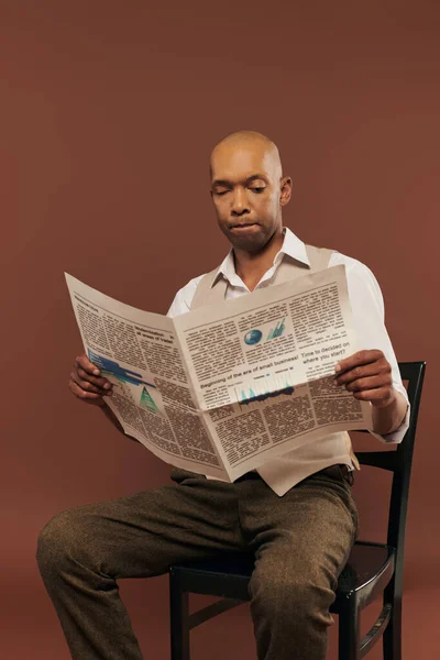Inclusion, bold african american man with myasthenia gravis syndrome sitting on chair and reading newspaper, dark skinned man with autoimmune disease on brown background — Stock Photo