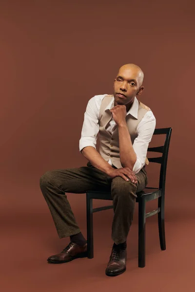 Inclusion, bold african american man with myasthenia gravis syndrome, sitting on chair and looking at camera, dark skinned man with chronic disease on brown background — Stock Photo