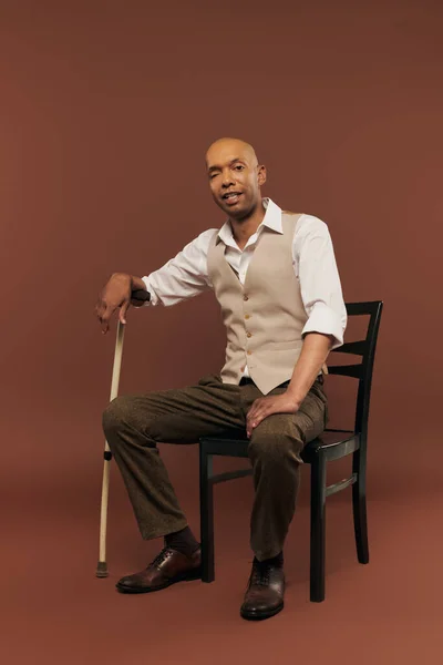Inclusion, real people, african american man with myasthenia gravis syndrome sitting on chair and looking at camera, dark skinned man with chronic disease holding walking stick on brown background — Stock Photo