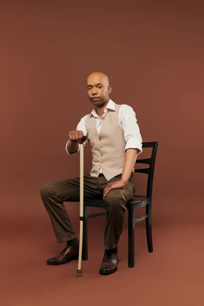 Inclusion, bold african american man with myasthenia gravis syndrome, sitting on chair and leaning on walking stick, looking at camera, dark skinned man with chronic disease on brown background — Stock Photo