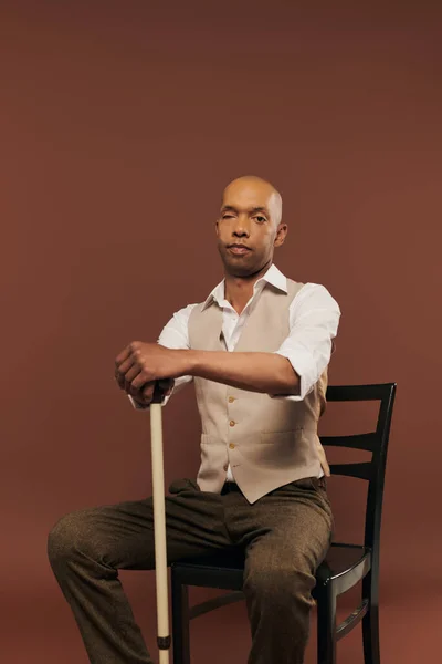 Inclusion, african american man with myasthenia gravis syndrome, sitting on chair and leaning on walking cane, looking at camera, bold dark skinned man with chronic disease on brown background — Stock Photo