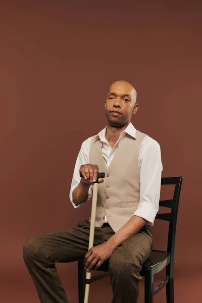 African american man with myasthenia gravis syndrome, sitting on chair and leaning on walking cane, looking at camera, bold dark skinned man with chronic disease on brown background, inclusion — Stock Photo