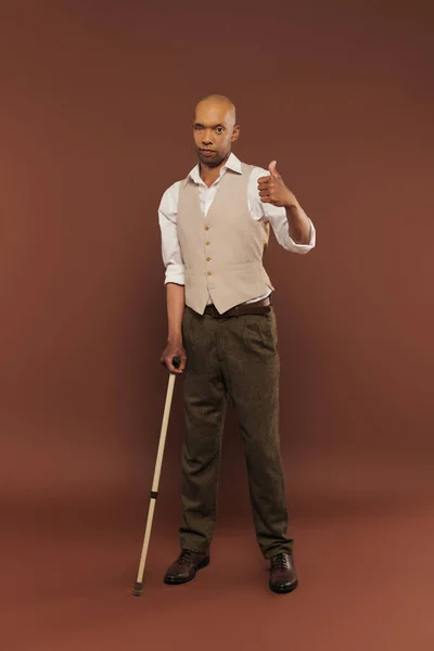 Homme afro-américain avec syndrome de myasthénie grave debout et appuyé sur la canne à pied, regardant la caméra, homme à la peau foncée audacieuse avec une maladie chronique montrant comme sur fond brun, inclusion — Photo de stock
