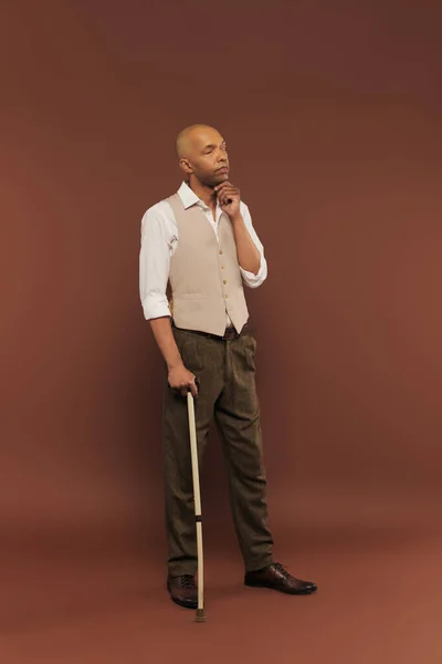 Inclusion, thoughtful african american man with myasthenia gravis syndrome leaning on walking cane, looking away, bold dark skinned man with chronic disease standing on brown background — Stock Photo