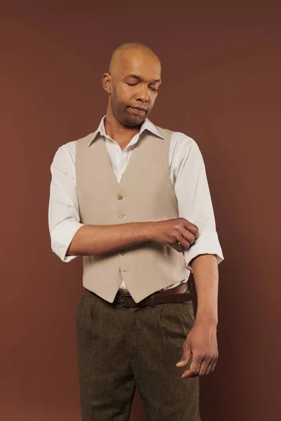 Bold african american man with myasthenia gravis standing on brown background, adjusting sleeve on white shirt, dark skinned, diversity and inclusion, real people, physical impairment — Stock Photo