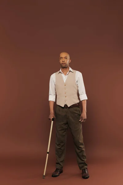Vraies personnes, homme afro-américain audacieux avec syndrome de myasthénie grave debout avec canne à pied sur fond brun, debout et regardant la caméra, la diversité et l'inclusion, déficience physique — Photo de stock