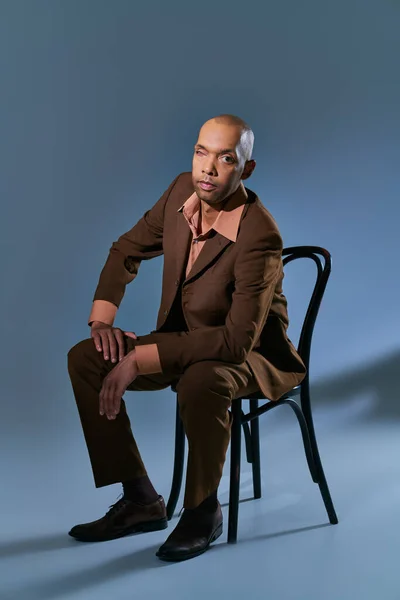 Homem negro afro-americano com síndrome de miastenia gravis sentado na cadeira e olhando para a câmera em fundo azul, pessoa de pele escura em terno, diversidade e inclusão, condição médica — Fotografia de Stock