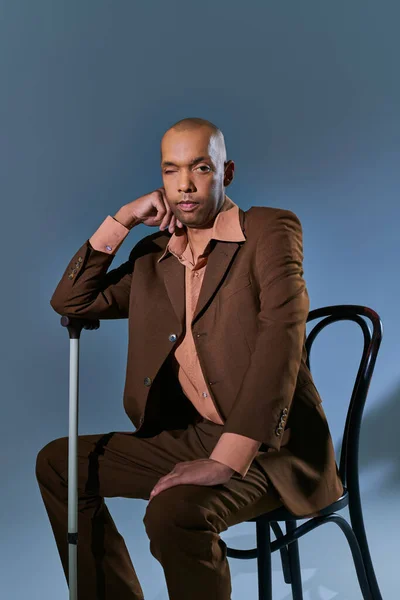 Diversity and inclusion, ptosis, african american man with myasthenia gravis syndrome sitting on chair and looking at camera on blue background, leaning on walking cane, difficulty walking — Stock Photo