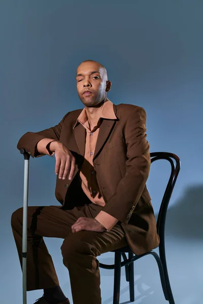 Разнообразие и включение, птоз, смелый африканский американец с синдромом миастении, сидящий на стуле и смотрящий на камеру на синем фоне, опирающийся на трость, трудности при ходьбе — стоковое фото