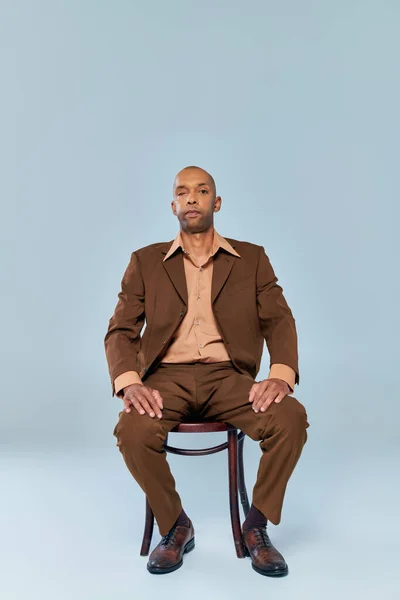 Real people, full length of bold african american man with myasthenia gravis sitting on wooden chair on grey background, dark skinned person in suit looking at camera, diversity and inclusion — Stock Photo
