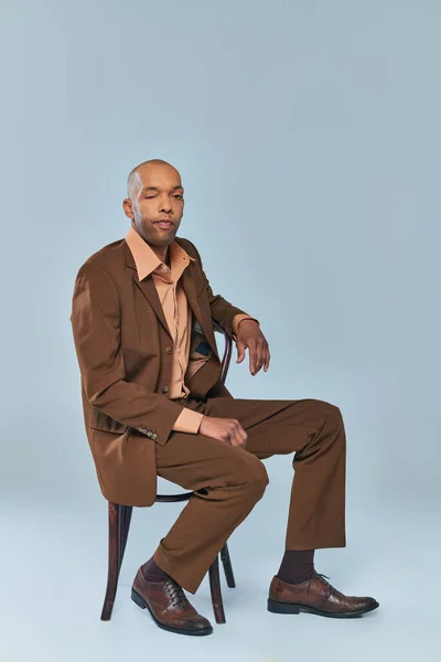 Handicap, pleine longueur de gras homme afro-américain avec myasthénie gravis assis sur une chaise en bois sur fond gris, personne à la peau foncée en costume regardant la caméra, la diversité et l'inclusion — Photo de stock