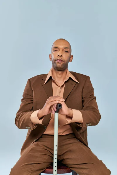 Disability, bold african american man with myasthenia gravis sitting on wooden chair on grey background, dark skinned person in suit leaning on walking cane, diversity and inclusion — Stock Photo