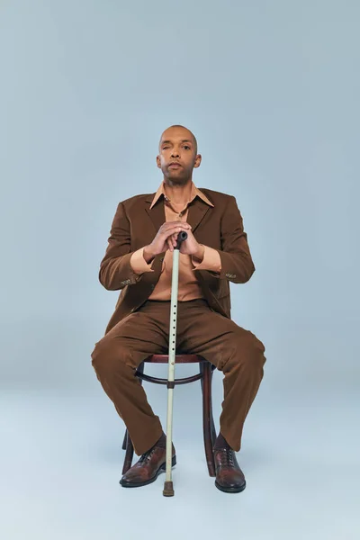 Disability, full length of bold african american man with myasthenia gravis syndrome sitting on chair on grey background, dark skinned person in suit leaning on walking cane, diversity and inclusion — Stock Photo