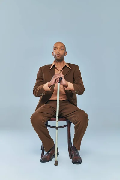 Handicap, pleine longueur de gras homme afro-américain avec le syndrome de la myasthénie grave assis sur une chaise sur fond gris, personne à la peau foncée en costume penché sur la canne à pied, la diversité et l'inclusion — Photo de stock