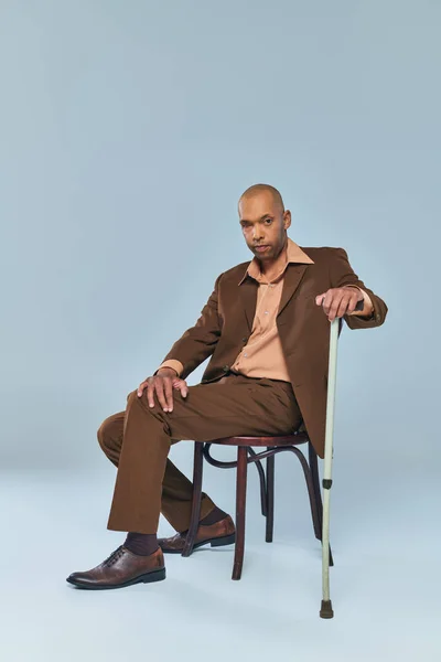 Етнічний, повнометражний сміливий афроамериканський чоловік з міастенією гравієм, що сидить на стільці на сірому фоні, темношкіра людина в костюмі, спираючись на прогулянкову тростину, різноманітність та включення — стокове фото