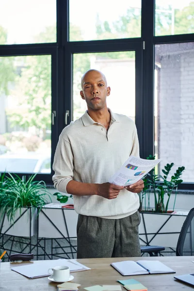 Miastenia, trabalhador de escritório de pele escura com síndrome de ptose segurando gráficos, inclusão, negrito empresário afro-americano com síndrome do olho olhando para a câmera, de pé perto de plantas verdes no escritório — Fotografia de Stock