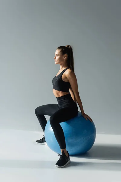 Sportswoman ajuste em sutiã de esportes preto e leggings sentado na bola de fitness em fundo cinza — Fotografia de Stock