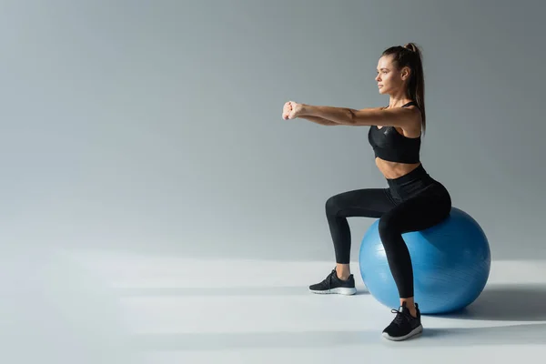 Fit brunette sportswoman in black active wear exercising on fitness ball on grey background, balance — Stock Photo
