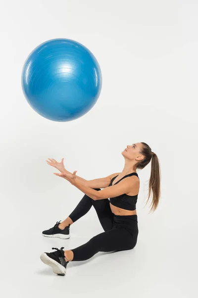 Woman in black sporty outfit throwing fitness ball and sitting on white background, athletic concept — Stock Photo