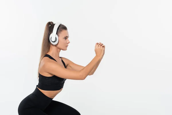 Sportlerin in schwarzer Aktivkleidung mit Kopfhörern bei Kniebeugen auf weißem Untergrund — Stockfoto
