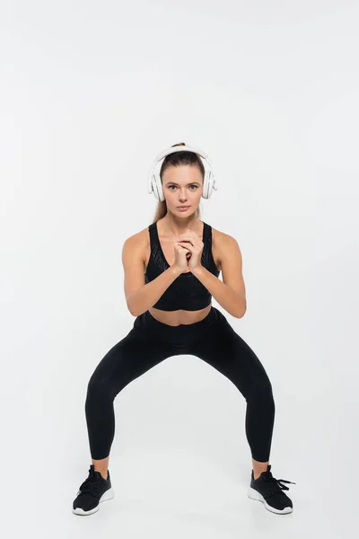 Fitte Sportlerin in Sportbekleidung mit Kopfhörern und Kniebeugen auf weißem Hintergrund — Stockfoto