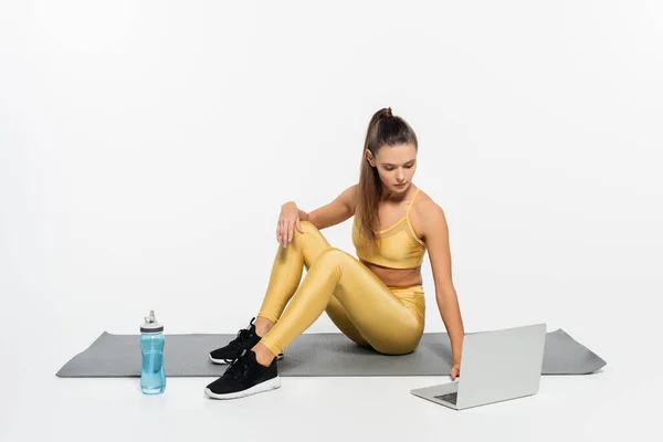 Woman in active clothes using laptop while sitting near bottle and fitness mat on white background — Stock Photo