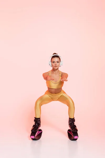 Endurance and balance, woman in kangoo jumping shoes and wireless headphones doing squats on pink — Stock Photo