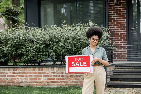 Heureux courtier immobilier afro-américain tenant enseigne avec à vendre lettrage près de maison en ville — Photo de stock
