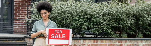 Positive courtier immobilier afro-américain debout avec à vendre enseigne près de bâtiment, bannière — Photo de stock