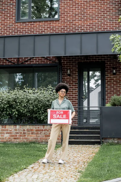 Pleased african american property agent holding signboard with for sale lettering near city house — Stock Photo