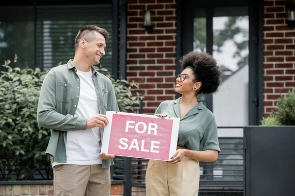 Happy multiethnic real estate brokers holding for sale signboard, looking at each other near house — Stock Photo