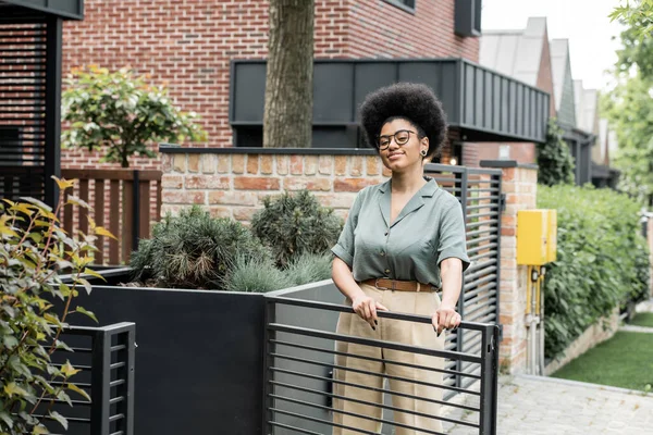 Agente inmobiliario afroamericano sonriendo cerca de valla y plantas verdes junto a casa urbana - foto de stock