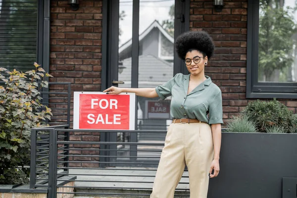 Agent immobilier afro-américain optimiste debout avec à vendre enseigne près de la clôture du chalet — Photo de stock