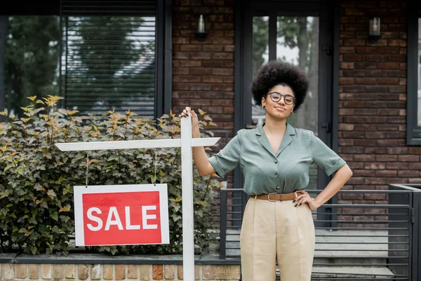 Afro-américain agent immobilier souriant à la caméra près de maison à vendre sur la rue de la ville — Photo de stock