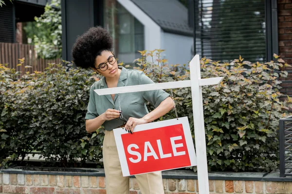 African american property agent hanging for sale signboard near house on city street — Stock Photo