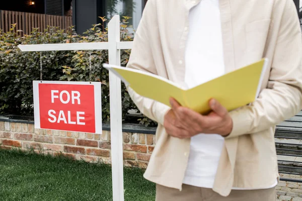Cropped view of property realtor standing with folder near for sale signboard in front of city house — Stock Photo